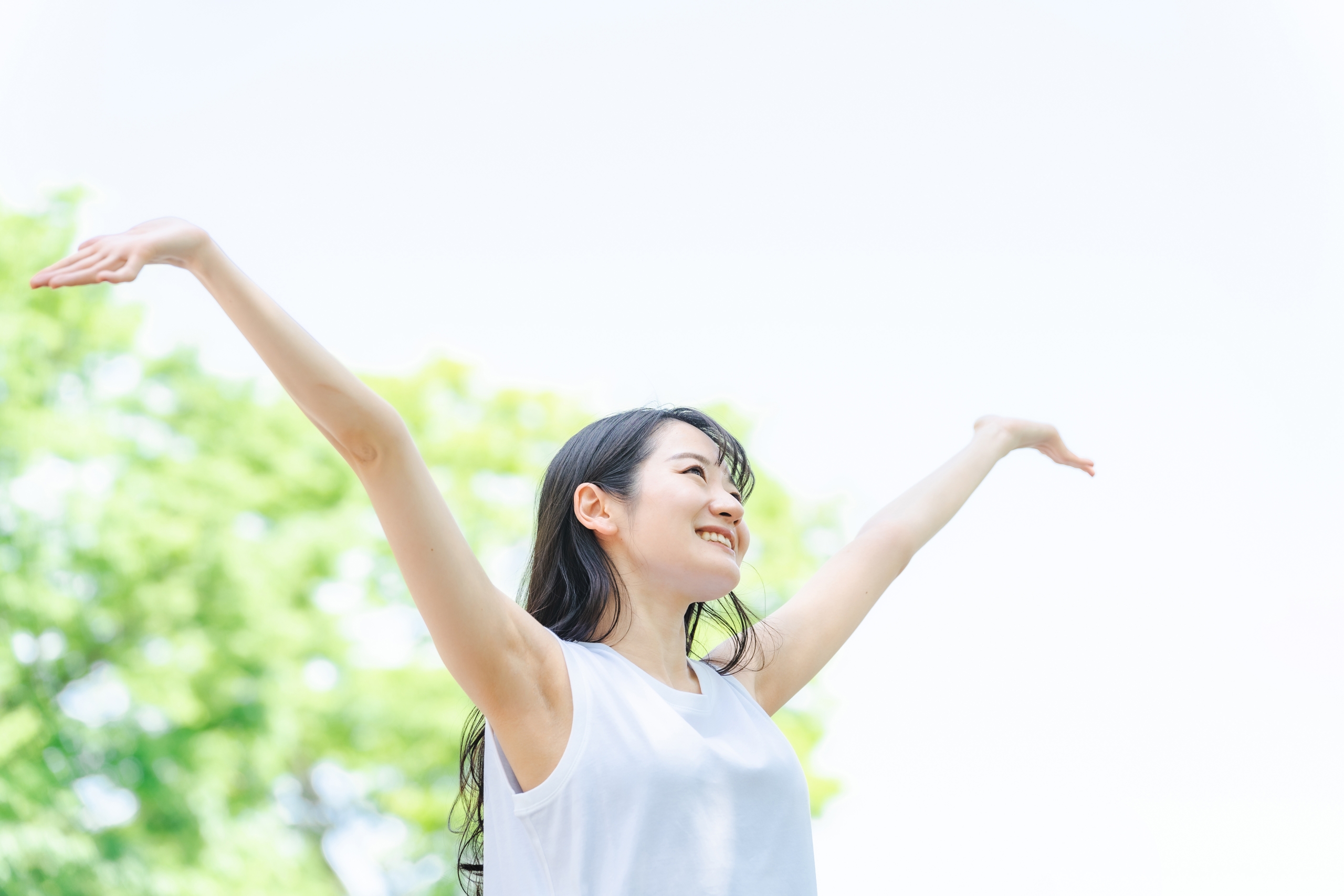 新緑の木々を背景に、白い服を着た若い女性が両手を大きく広げ、気持ちよさそうに空を見上げている。リフレッシュ、解放感、健康的なライフスタイルを象徴する場面。
